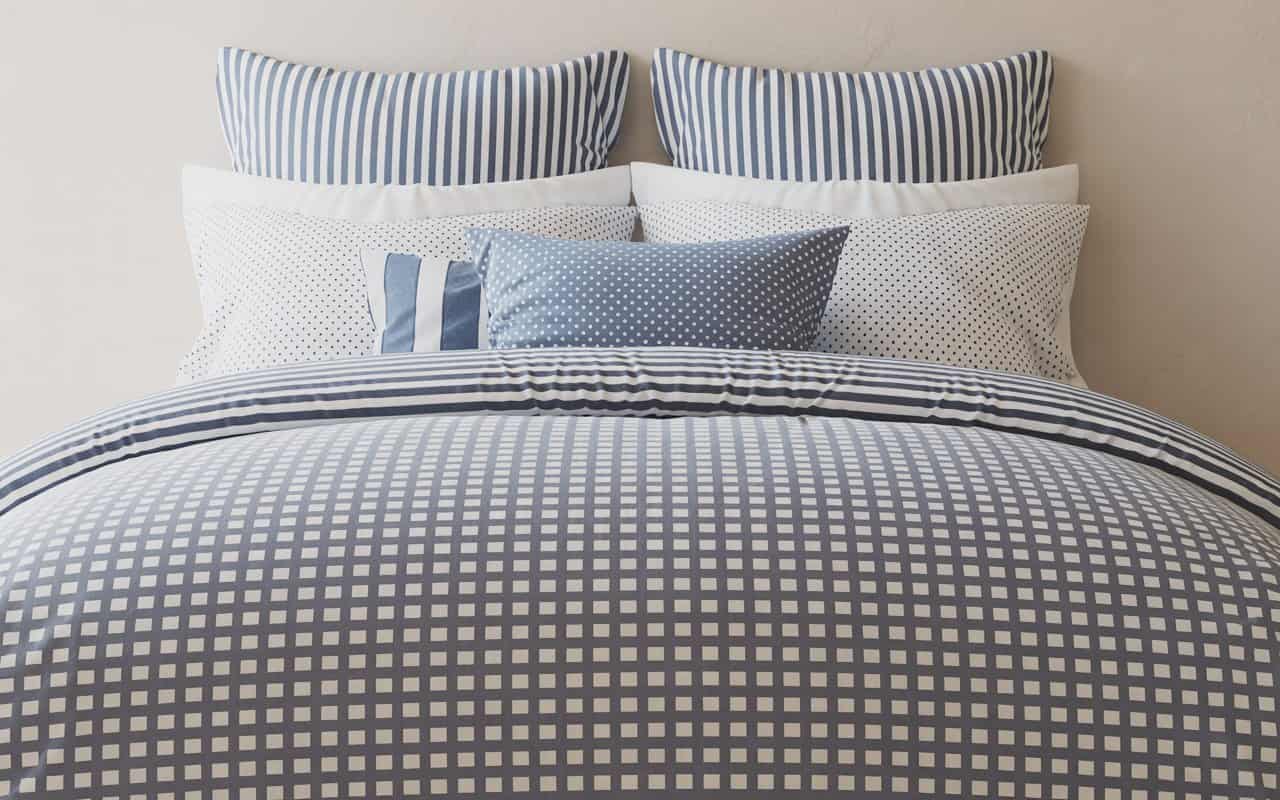 A bed styled with layered patterns. A large-scale grey checkered duvet, medium-scale striped shams, and small-scale polka dot accent pillows, all in a blue and white color palette.