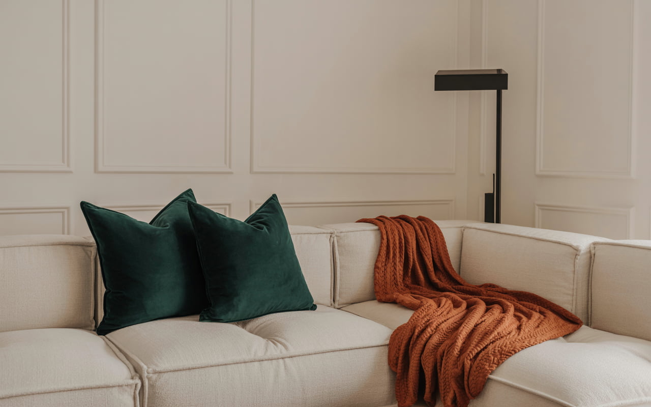 A sophisticated living room with off-white walls. A large sand-colored sectional sofa is decorated with two deep emerald green velvet pillows and a burnt orange knit throw. A minimalist black floor lamp stands in the corner.