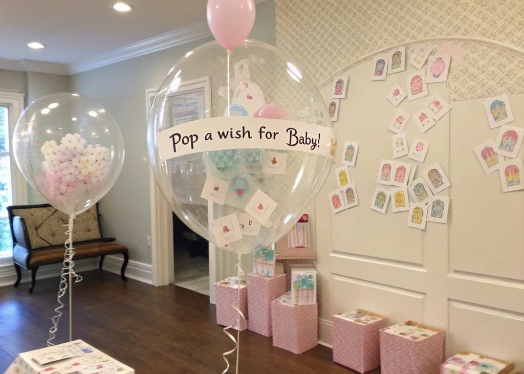 “Clear balloons filled with pastel cards inside, arranged around a baby shower room, with a sign saying ‘Pop a Wish for Baby!’”