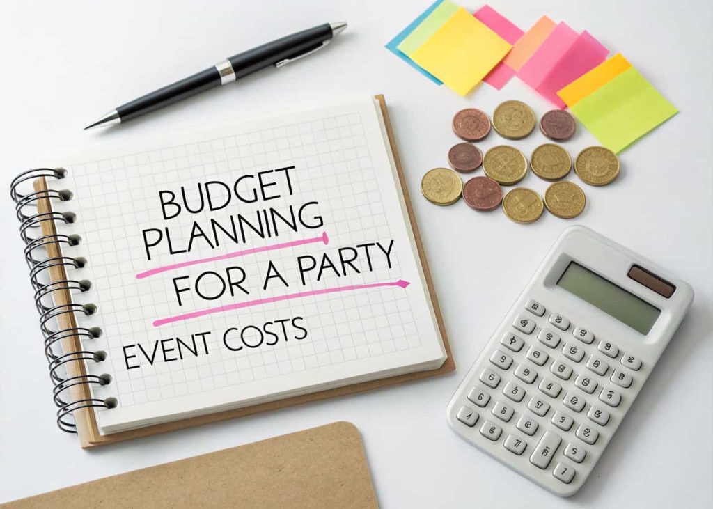 A flat lay of budget planning for a party: notebook, calculator, coins, and colorful sticky notes labeled with event costs."