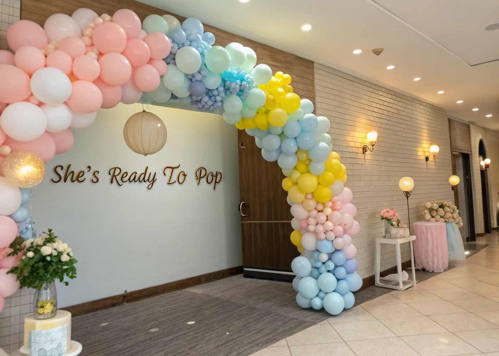 A vibrant balloon garland in pastel pink, blue, yellow, and white forming an arch at a baby shower entrance, with soft lighting and cute “She’s Ready to Pop” signs.