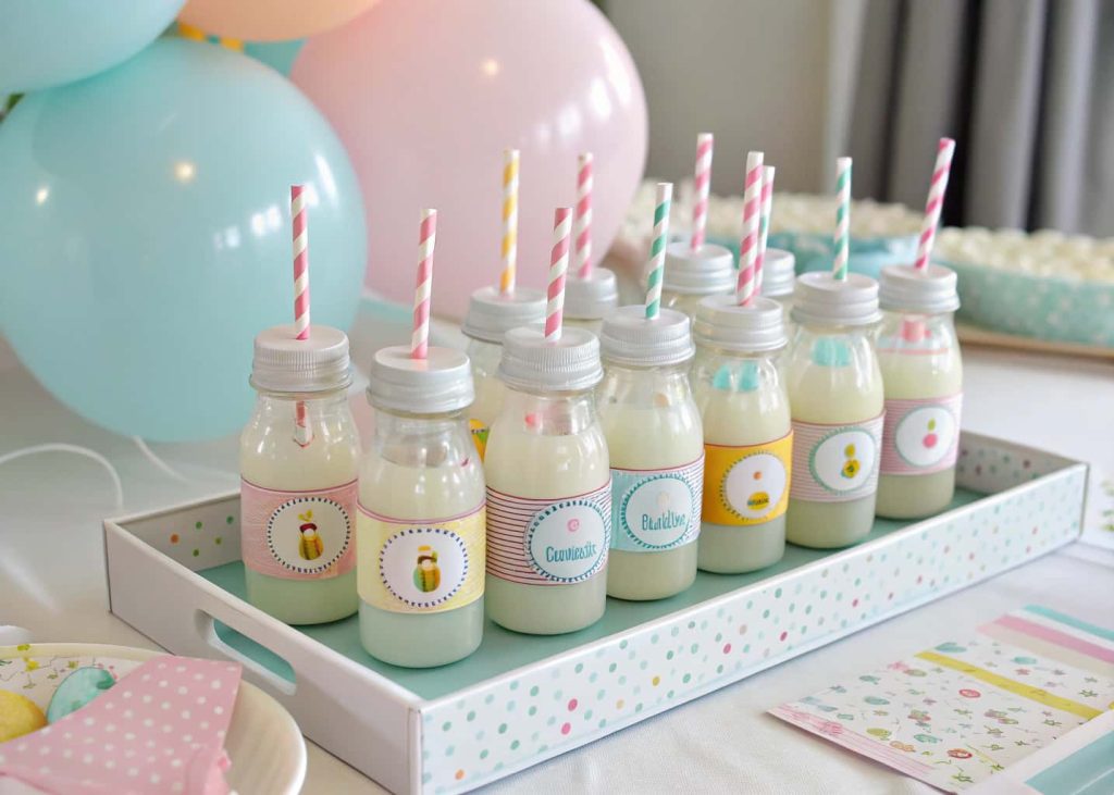 A baby shower drink station with pastel-colored baby bottles, fun labels, straws, and balloons around the tray.”