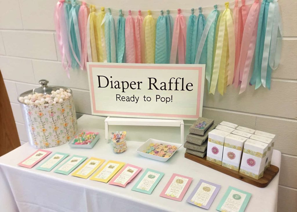 A baby shower diaper raffle table with pastel ribbons, tickets, small prizes, and a sign saying ‘Diaper Raffle – Ready to Pop!’”