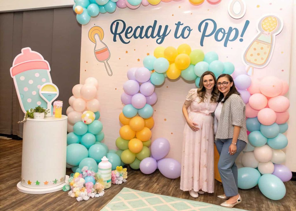 “A baby shower photo booth with pastel balloon backdrop, baby-themed props like rattles and bottles, and a ‘Ready to Pop!’ sign.”
