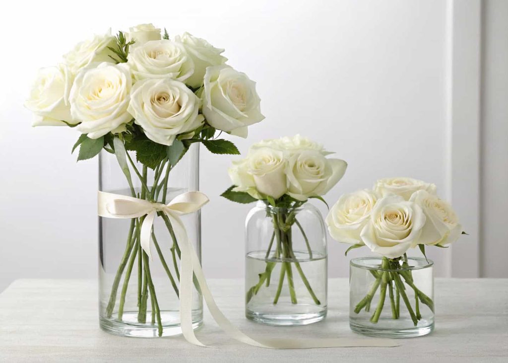 "Minimalist bloom bar with white roses, clear glass vases, simple ribbons, modern clean setup."