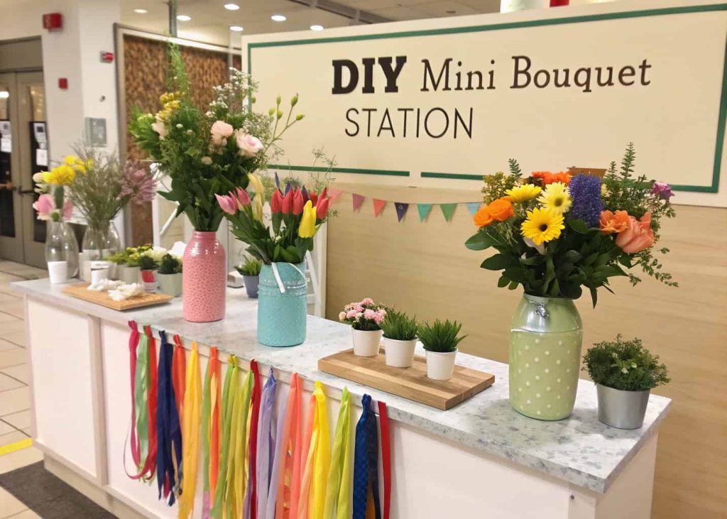 "DIY mini bouquet station with assorted flowers, small vases, ribbons, instructional signage, bright event setup."