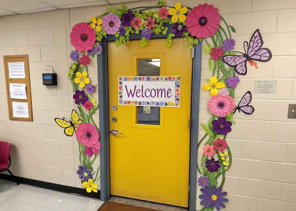 A classroom door decorated with a colorful floral wreath, butterflies, and a “Welcome” sign in the center.