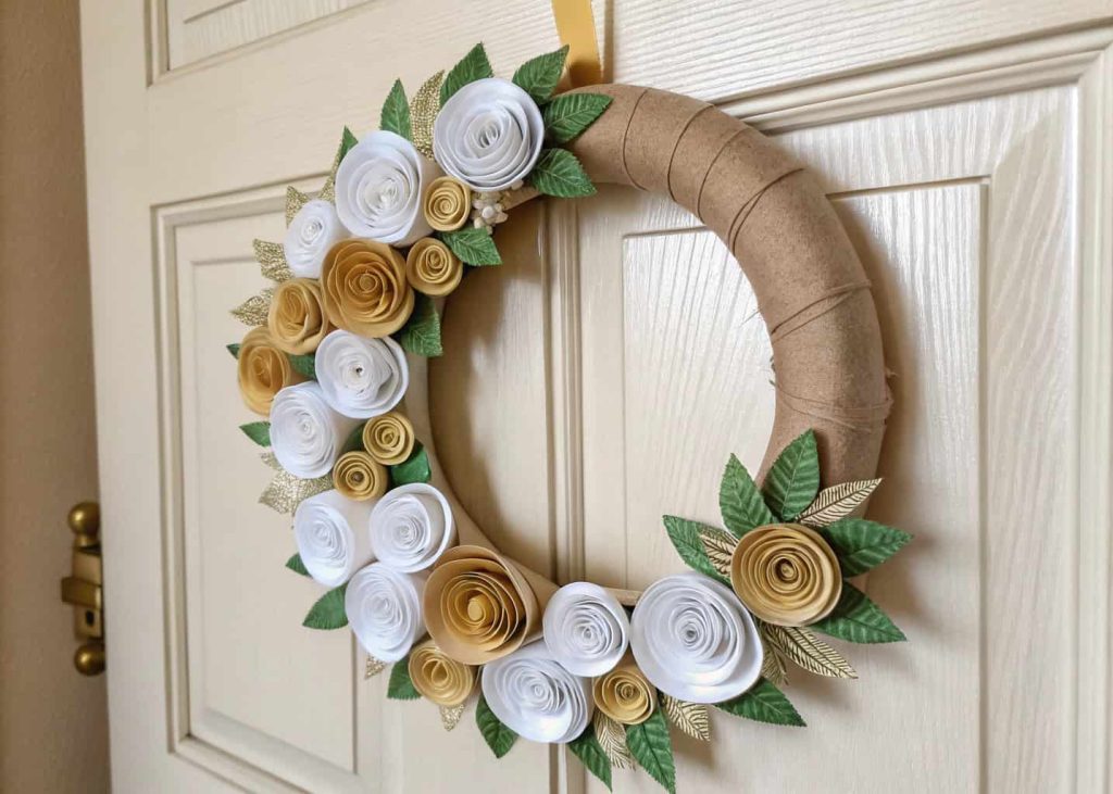 Handmade paper wreath with white, gold, and green rolled paper flowers hanging on a light wooden door.