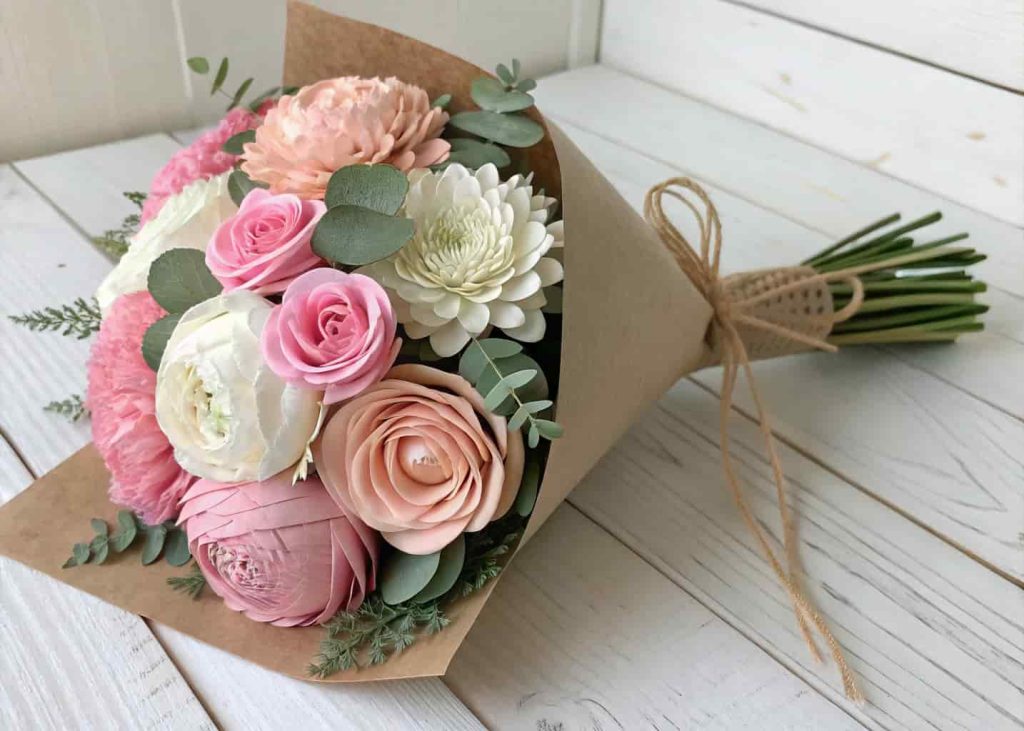A bouquet of handmade paper peonies and roses in pink and white tones wrapped in kraft paper on a wooden surface.