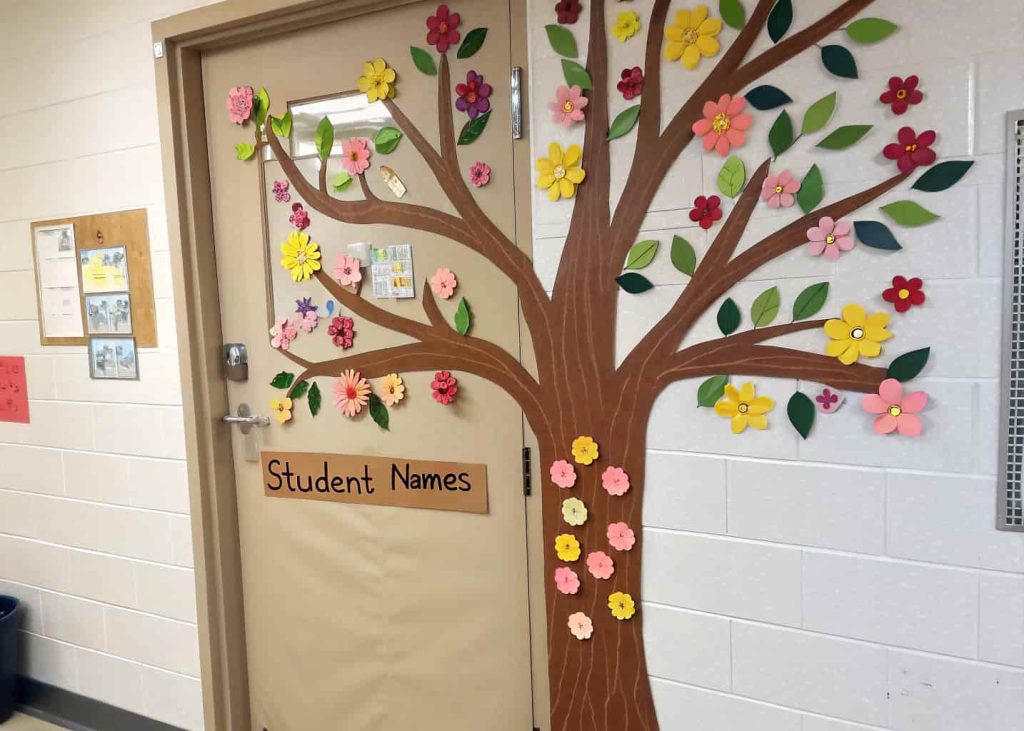 Classroom door with a brown paper tree, colorful flowers and leaves, some labeled with student names.
