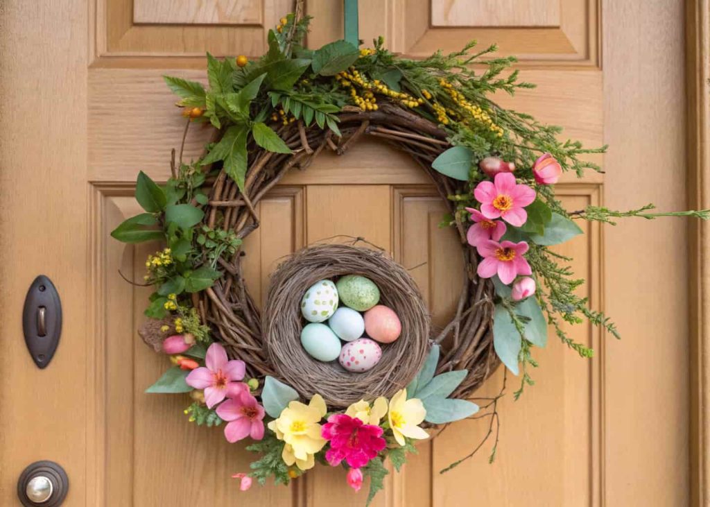 A spring wreath with a small bird nest, pastel eggs, and flowers hanging on a wooden front door.