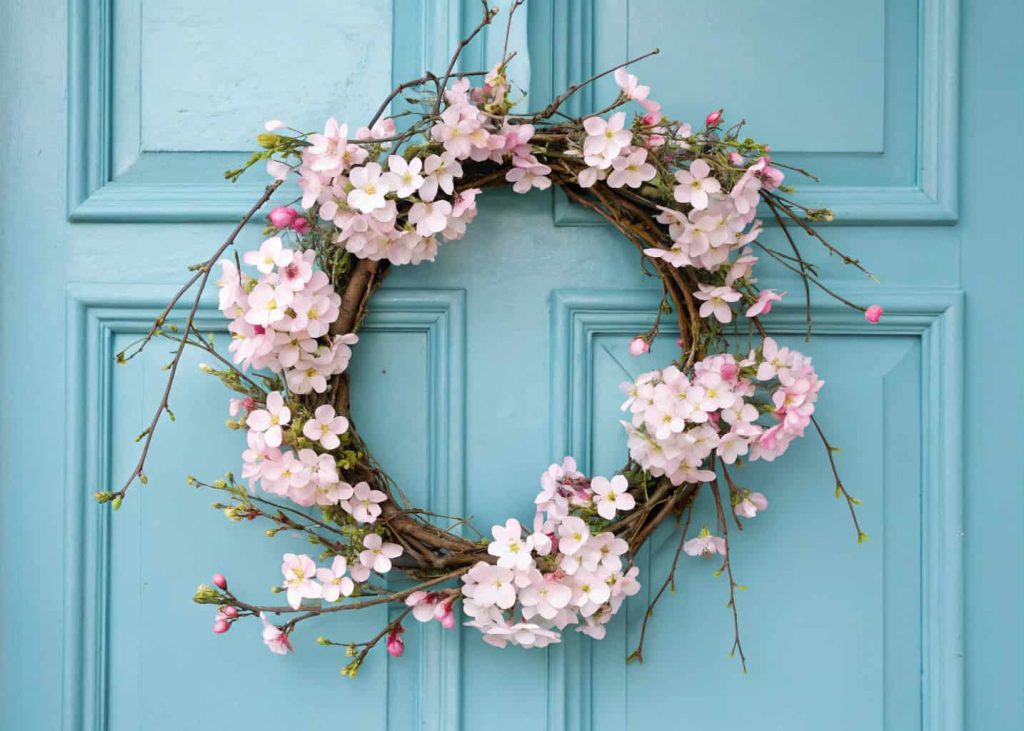 A wreath with soft pink cherry blossom branches and white flowers hanging on a pastel blue front door.