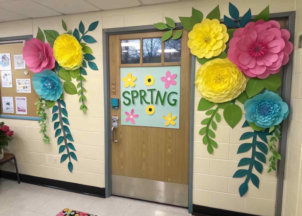 A classroom door covered with large, colorful paper flowers in pink, yellow, and blue with green leaves, a cheerful spring sign in the center.