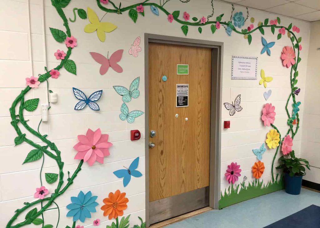 Classroom door decorated with multi-colored butterflies flying upward, surrounded by paper flowers and green vines, playful and lively spring theme.