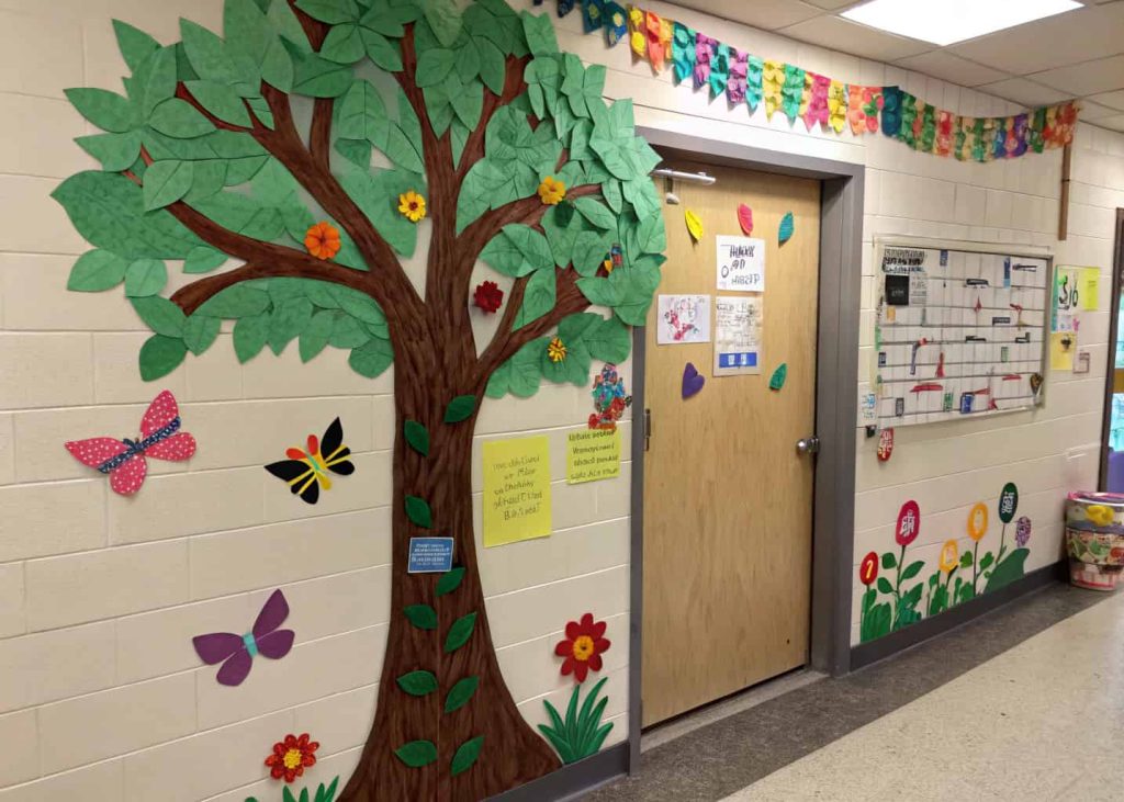 Door with a large paper tree, green leaves, colorful flowers, butterflies, and student artwork hanging like ornaments.