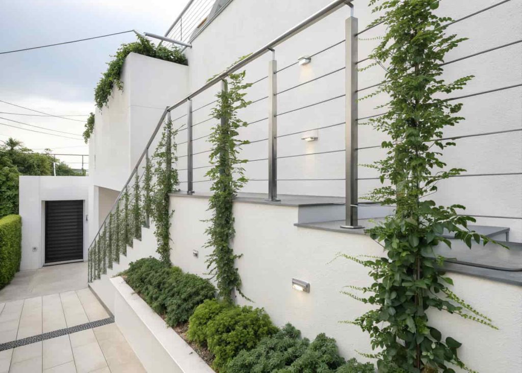 Stainless steel horizontal wire cables mounted on a white modern building wall with lush green climbing plants, clean minimalist aesthetic, urban garden, sharp professional photography.