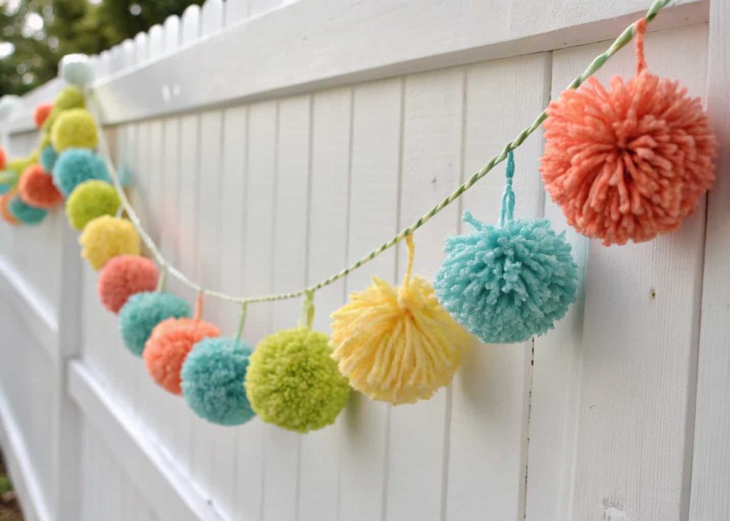 Fluffy colorful yarn pom pom garland in coral, yellow, sky blue, and green, hung across a white wall, cheerful spring party decor, bright natural daylight, festive and playful handmade aesthetic, close-up texture shot.