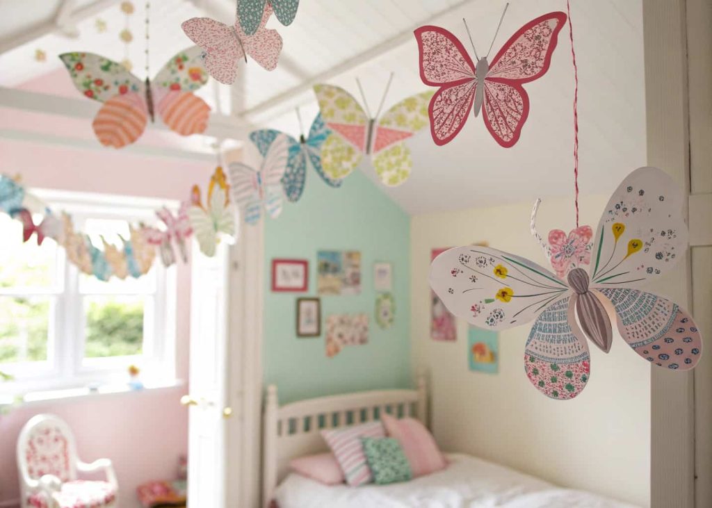 Whimsical paper butterfly garland in colorful and patterned cardstock, hung across a child's room ceiling, bright spring morning light, magical and playful storybook aesthetic, butterflies in various sizes and patterns, airy and joyful atmosphere.