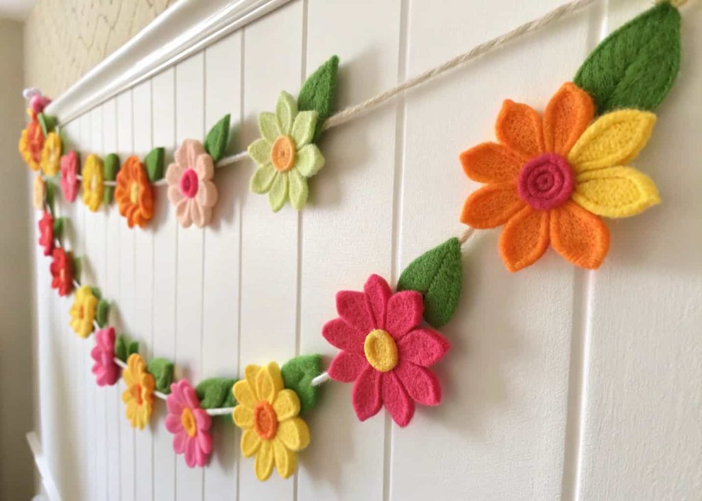 Bright felt flower garland in spring colors — pink, yellow, orange, and green — threaded on a thick cream yarn, hung on a white wall, close-up handmade texture shot, cheerful children's room or party backdrop decor, soft natural daylight.