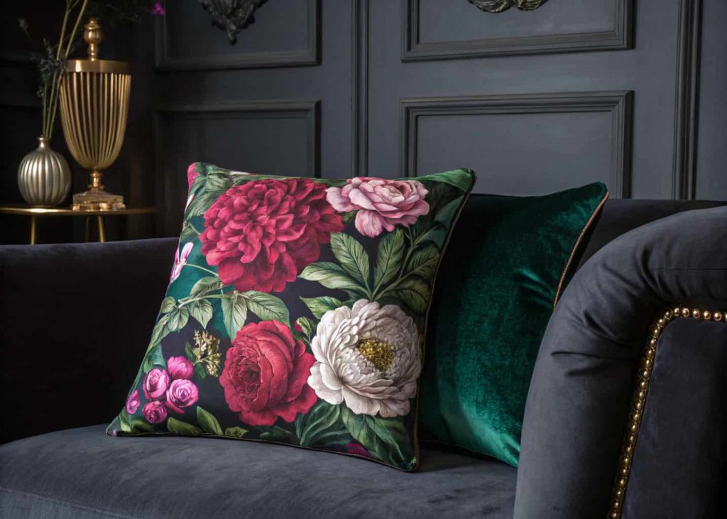 A large maximalist floral throw pillow with oversized peonies and roses in deep jewel tones, emerald green, burgundy, fuchsia, placed on a dark charcoal sofa, moody dramatic interior, editorial style photography