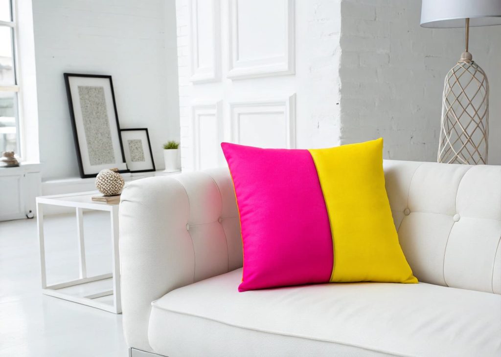  A bold neon color block throw pillow split into hot pink and electric yellow halves, placed on a white minimalist sofa, bright white room, modern art aesthetic, graphic design inspired interior, clean studio photography