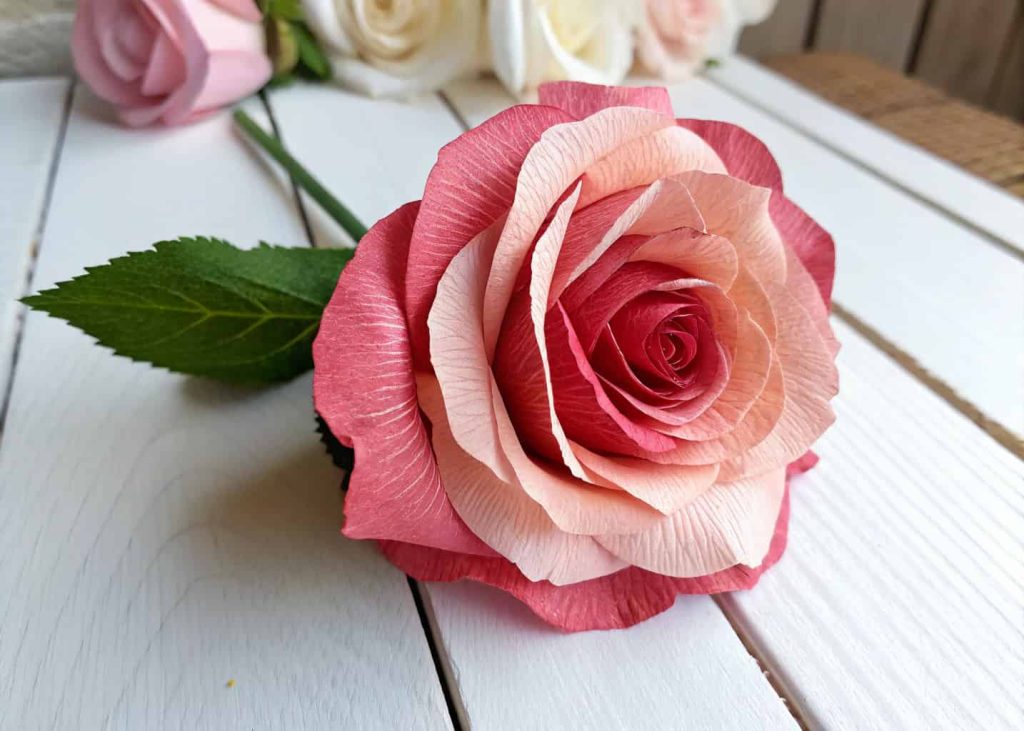 A beautiful handmade crepe paper rose in soft pink and red tones, placed on a white wooden surface with natural daylight, close-up macro shot, realistic craft photography style.