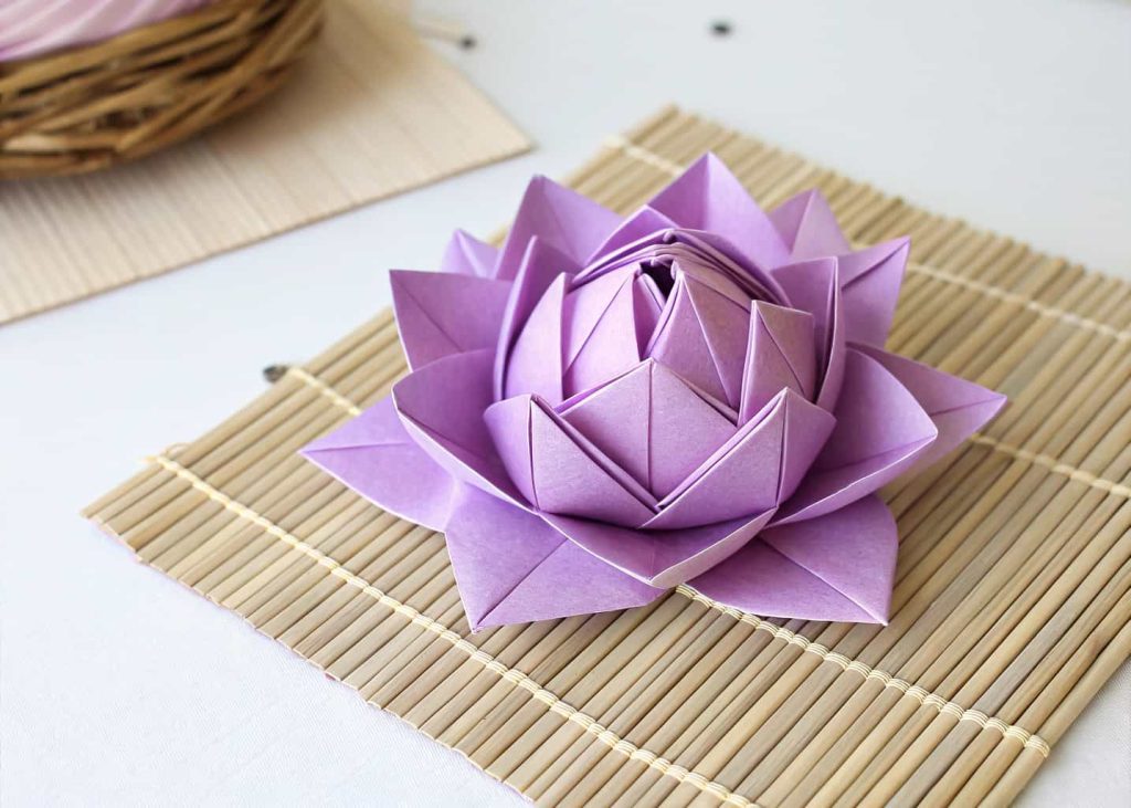 A neatly folded origami lotus flower made from pastel purple square paper, displayed on a bamboo mat, soft studio lighting, top-down flat lay photography, clean minimalist background.
