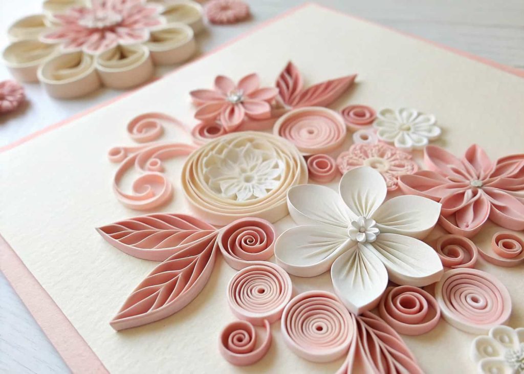 Detailed quilling paper flowers in blush pink and white arranged on a cream cardstock background, close-up photography showing the rolled paper texture, elegant and artistic craft style.