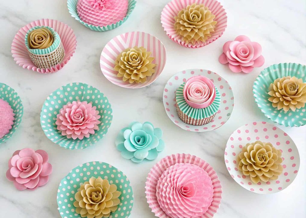 A collection of cupcake liner flowers in various polka dot and striped patterns, pink, mint, and gold, arranged on a white marble surface, overhead flat lay, bright airy photography for a baking blog.
