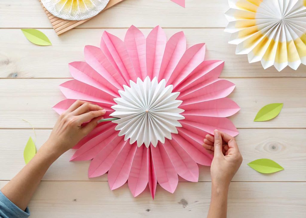 A colorful accordion-fold tissue paper flower in pink and white, hands gently pulling apart layers, soft natural light, flat lay on a wooden surface, bright and cheerful craft aesthetic.