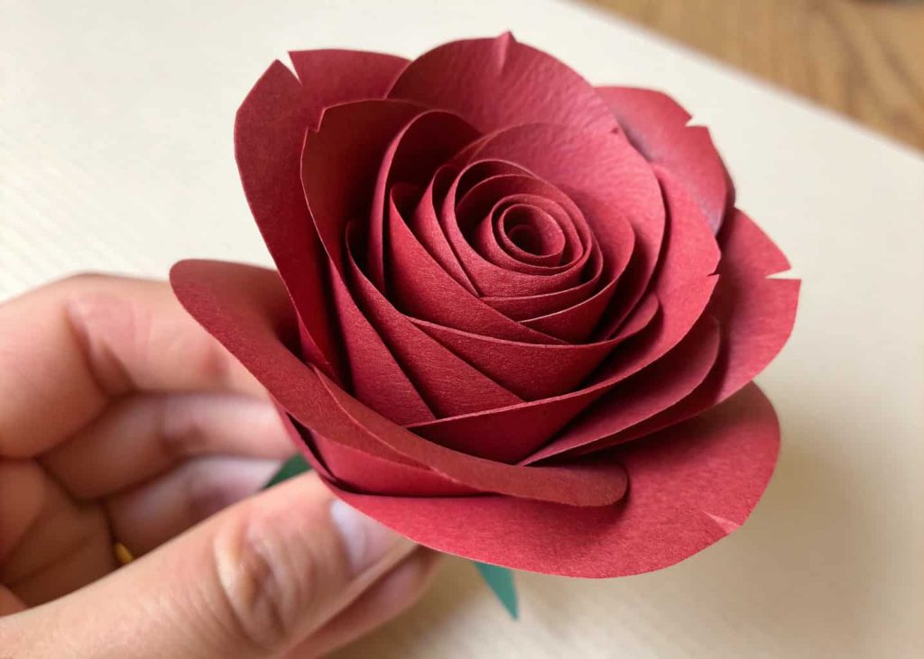 A handmade tissue paper rose in deep red, rolled petals with natural texture, held in a hand against a light beige background, close-up macro shot, romantic and artistic mood.