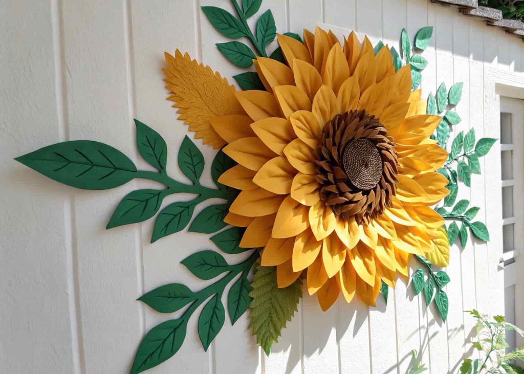 A large handmade tissue paper sunflower in golden yellow with a brown center, displayed on a white wall, surrounded by green paper leaves, vibrant and cheerful, bright daylight photography.