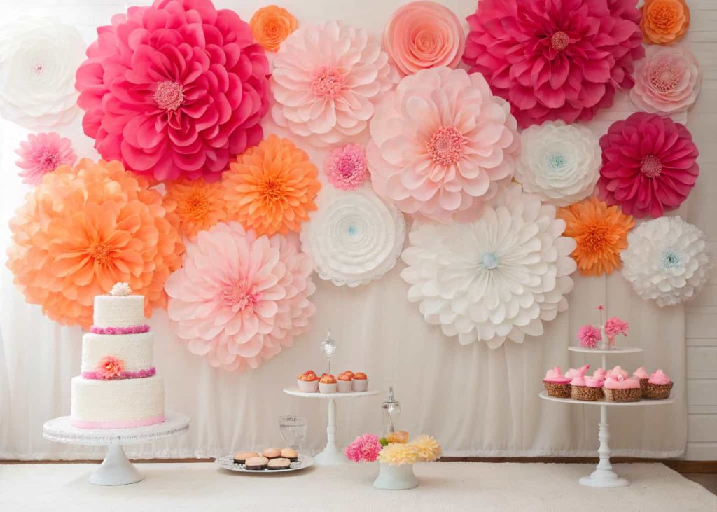 A colorful wall of giant tissue paper flowers in pink, coral, and white, used as a birthday party backdrop, hanging at different heights, vibrant and festive, professional event photography.