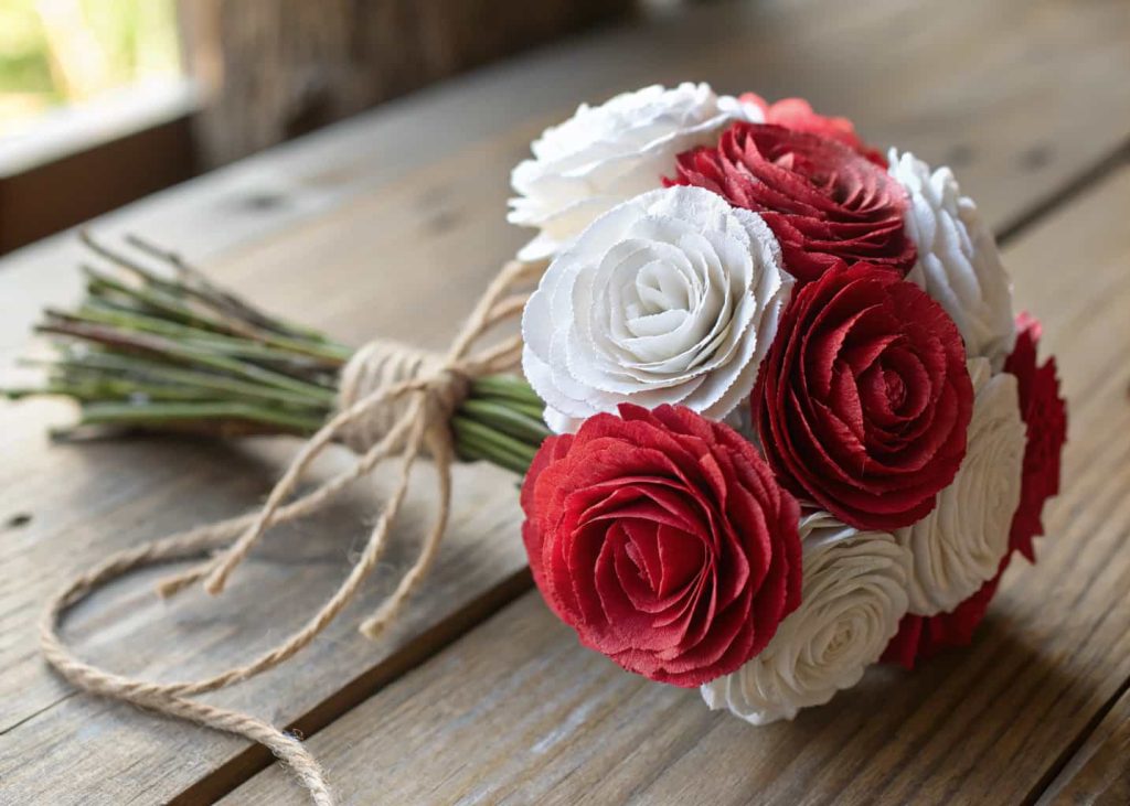 Handmade tissue paper carnations in red and white, bundled in a small bouquet, tied with twine, placed on a rustic wooden table, warm natural light, close-up craft photography.