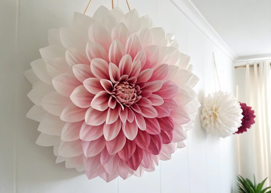 A large ombre tissue paper flower transitioning from deep rose to pale blush to white, hanging on a white wall, dreamy and romantic, soft studio lighting, high-quality craft photography.