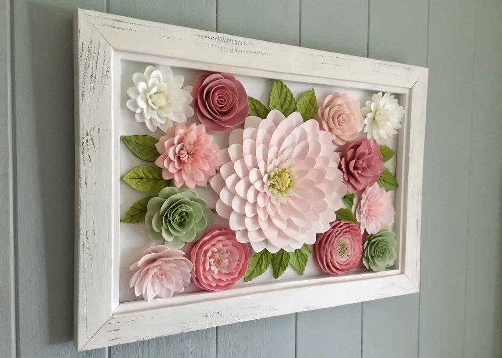 A framed wall art piece with tissue paper flowers in pink, white, and green arranged inside a white wooden frame, hung on a light gray wall, elegant home decor photography, soft lighting.