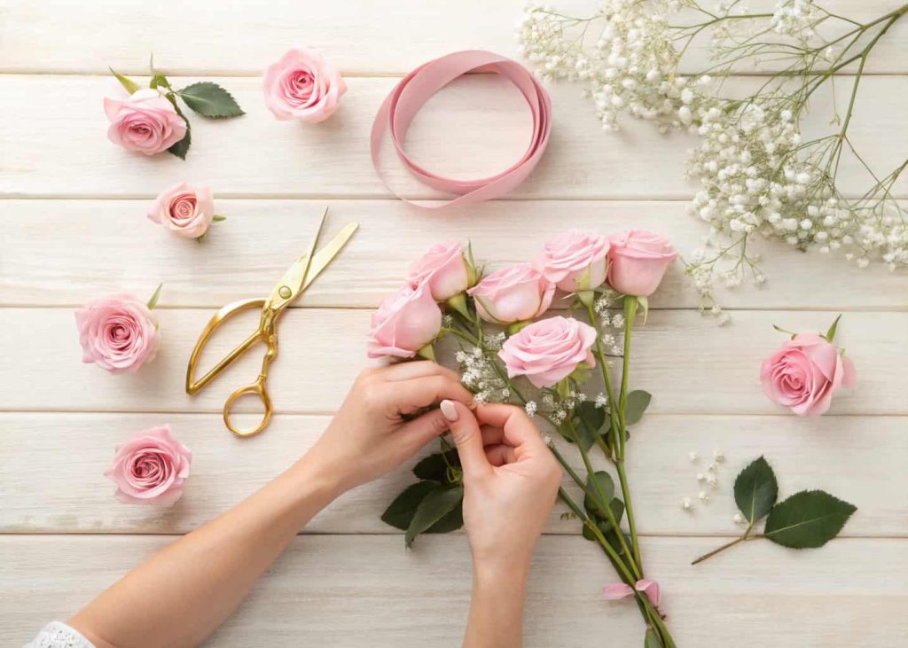Hands using gold scissors to trim flower stems on a wooden craft table, fresh pink roses and baby's breath nearby, flat lay perspective, natural daylight, soft feminine aesthetic