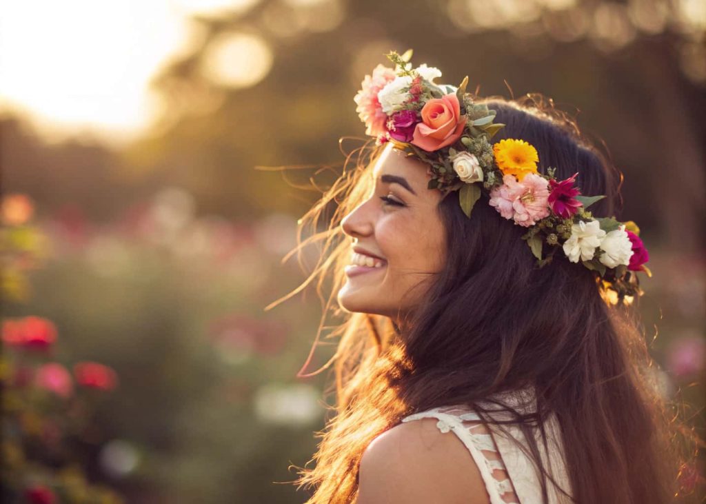 A young woman with flowing dark hair wearing a colorful flower crown outdoors, smiling naturally, golden hour sunlight, bokeh garden background, lifestyle photography, warm tones