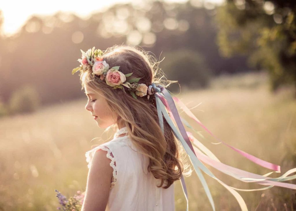 A flower crown with flowing pastel satin ribbons hanging from the back, worn by a girl in a sunny outdoor field, soft bokeh background, dreamy boho aesthetic photography.