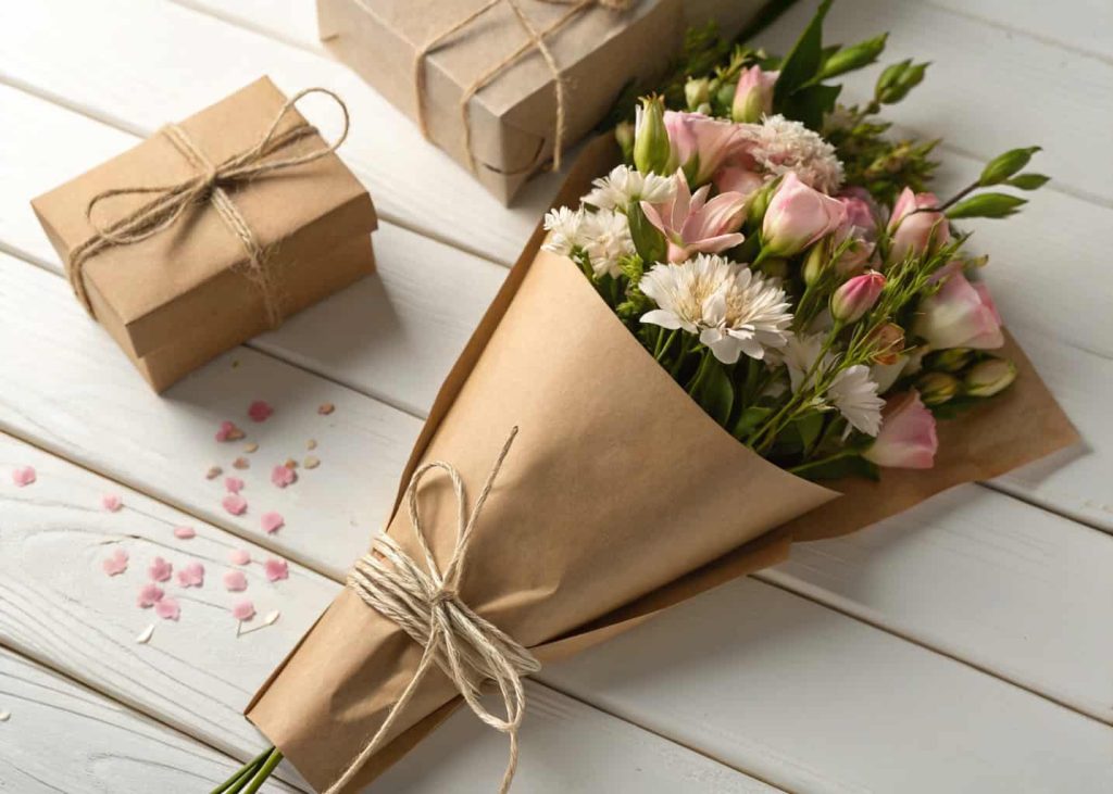 A flower bouquet wrapped in brown kraft paper and tied with natural twine sitting on a white wooden surface, soft romantic lighting, minimalist and elegant presentation, close-up shot.