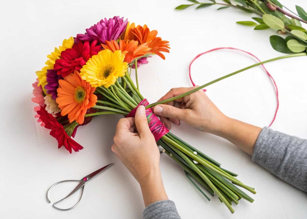 Close-up of hands using the spiral technique to hand-tie a colorful flower bouquet, stems being held at a diagonal angle, flowers fanning out beautifully, step-by-step tutorial style photography, white background.