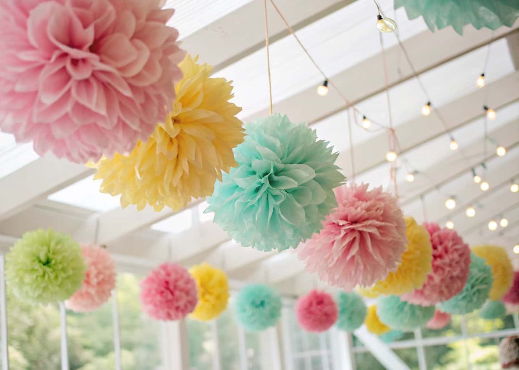 A vibrant display of colorful tissue paper pom-pom flowers hanging from a white ceiling with soft natural light, pink, yellow, and mint green colors, bokeh background, party decoration style, warm and cheerful atmosphere.