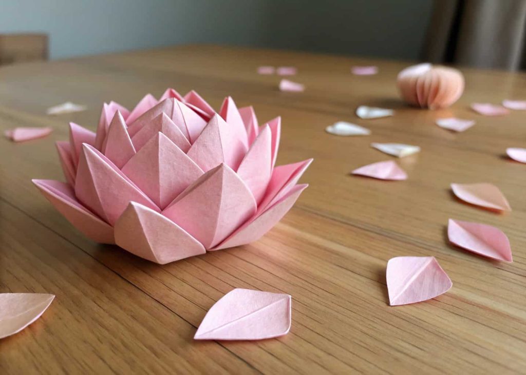  A delicate origami lotus flower made from light pink square paper, placed on a smooth wooden surface with a few scattered petals, soft ambient lighting, minimalist Japanese aesthetic, macro photography style.