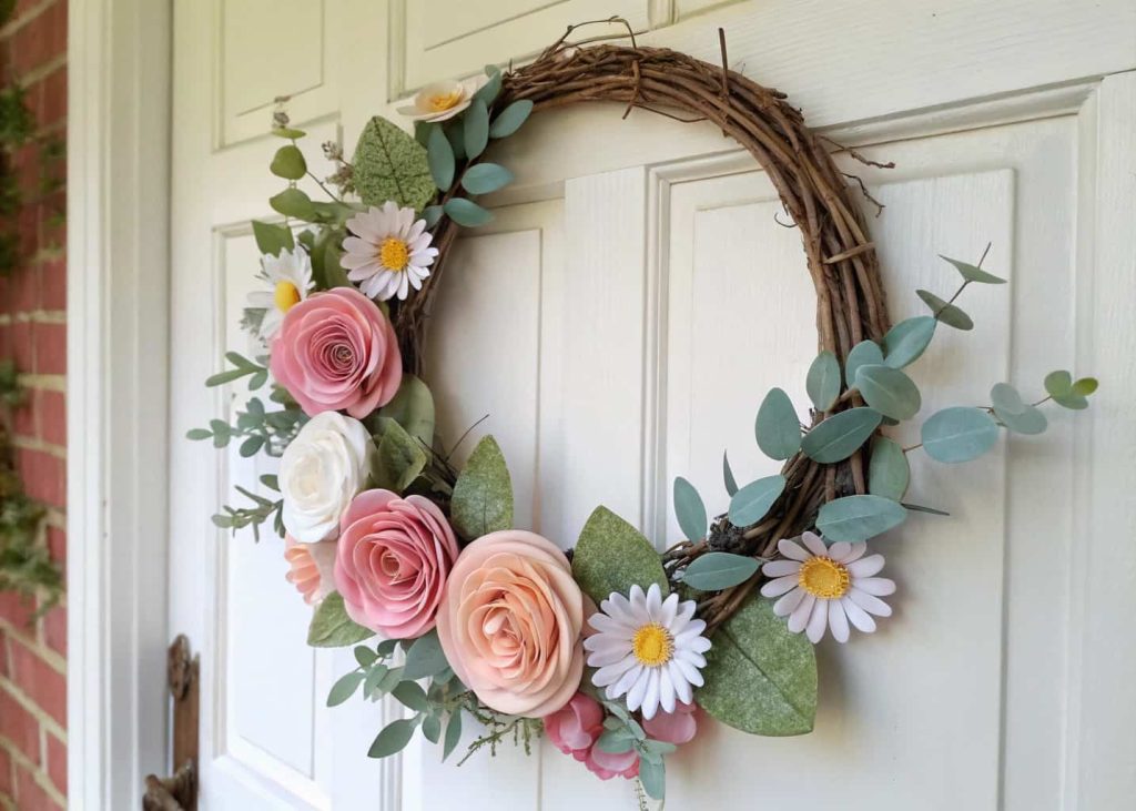 A beautiful handmade paper flower wreath featuring pastel roses, daisies, and eucalyptus leaves arranged on a circular vine base, hanging on a white rustic wooden door, spring morning natural light, charming and inviting home entrance aesthetic.