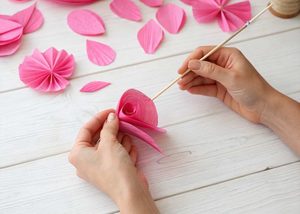 Hands gently curling the edge of a pink crepe paper petal around a wooden skewer, with several already-curled petals laying softly in the foreground on a light wooden surface.