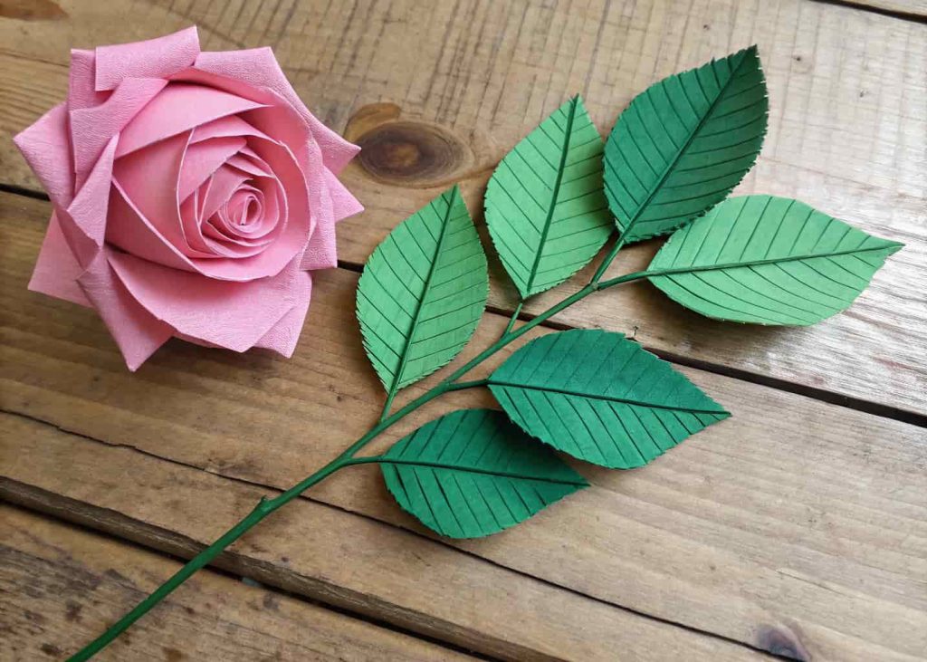 Handmade green crepe paper leaves with visible vein details attached to a flower stem with floral tape, placed next to a pink paper rose on a rustic wooden surface.
