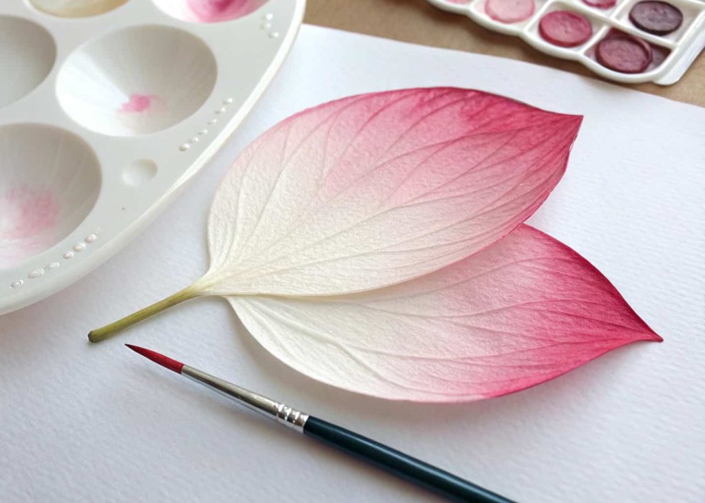 A watercolor-painted paper flower petal showing a soft ombre gradient from deep rose pink at the tip to pale blush white at the base, drying on a white ceramic palette with a fine brush nearby.