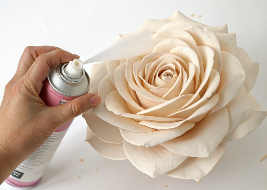 A finished handmade paper rose being lightly sprayed with a clear matte sealer from an aerosol can, held against a plain white background with a soft studio light highlighting the delicate petal texture.