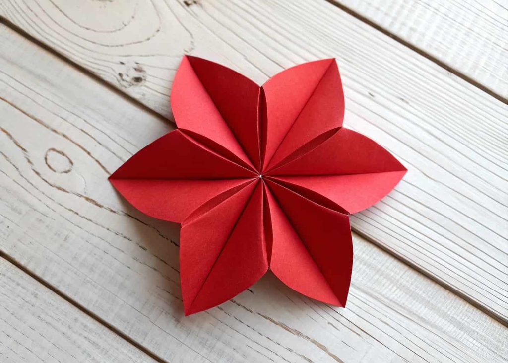 A cheerful 5-petal red paper flower made from a folded square sheet, lying flat on a white wooden table, soft natural daylight, top-down view, warm tones, minimalist craft aesthetic.