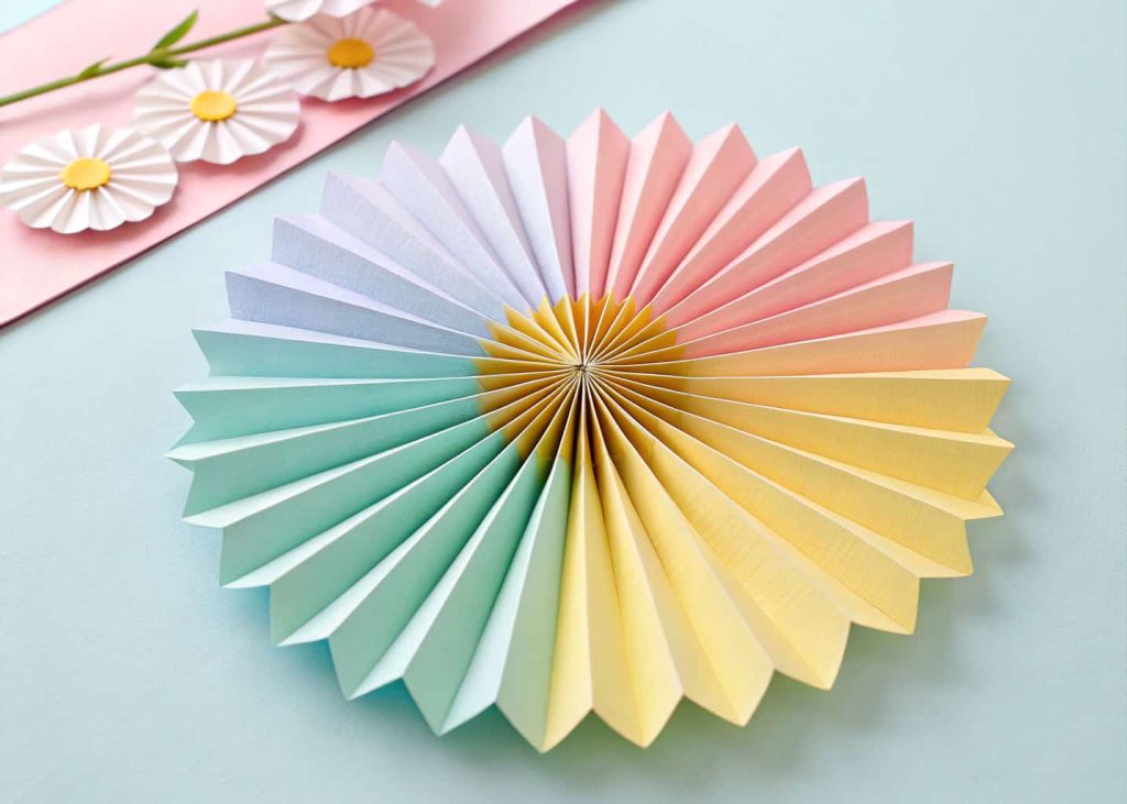 A colorful accordion fan paper flower made from pastel-colored sheets, pinched in the center with a yellow strip, placed on a light pastel background, airy and cheerful, soft studio lighting.